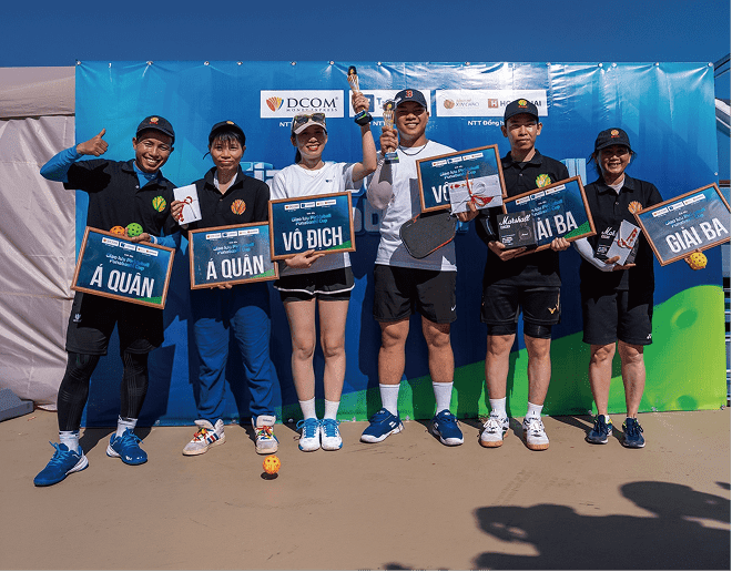 スポーツ大会の表彰式。青いパネルの前で、優勝（VÔ ĐỊCH）、準優勝（Á QUÂN）、3位（GIẢI BA）の盾やトロフィー、賞品を掲げて笑顔で並ぶ男女6名の選手たち。
